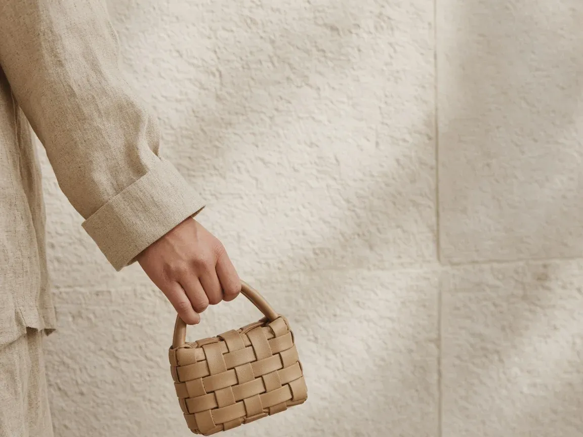A hand in a linen sleeve holding a small woven leather bag against a pale wall